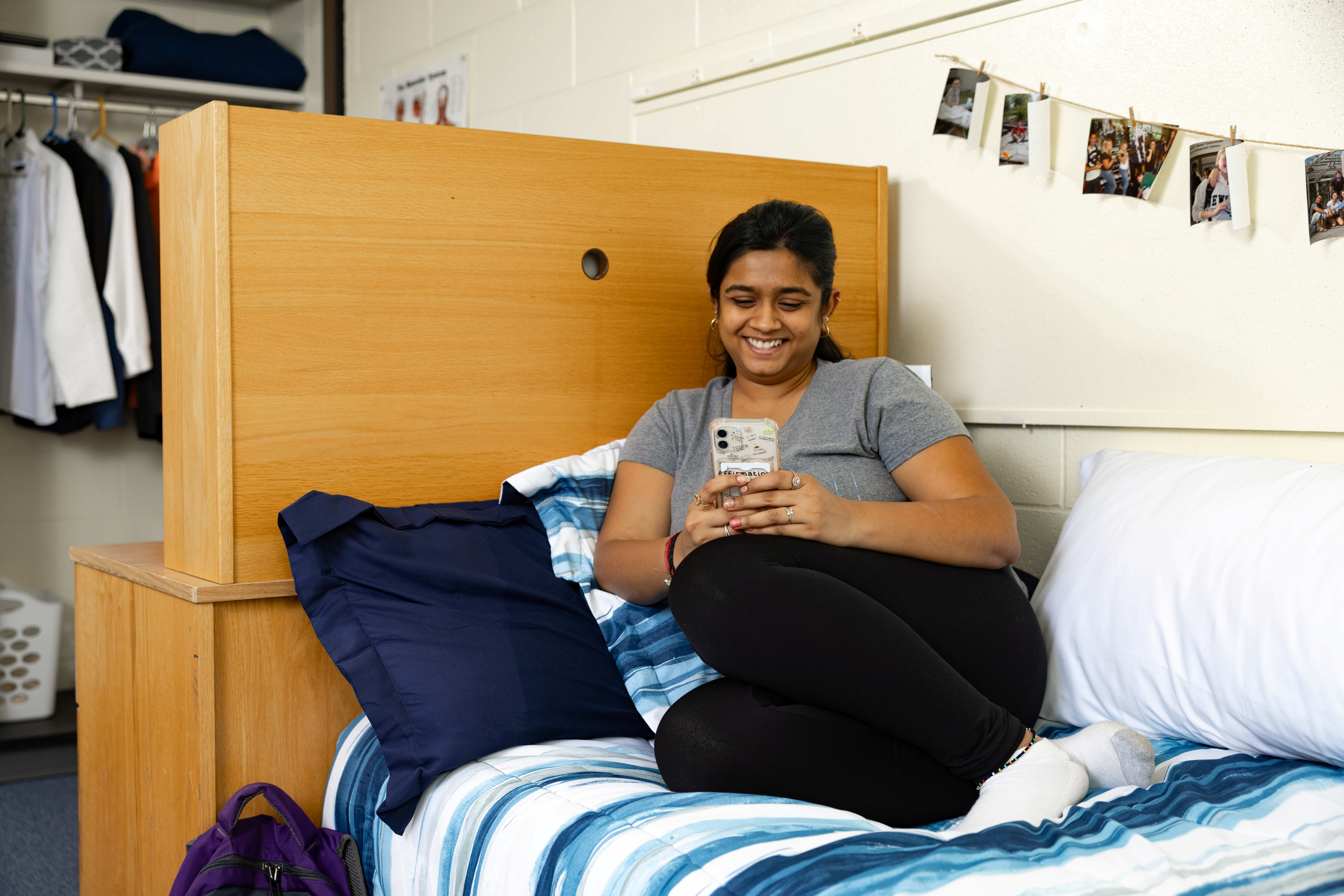 A college student smiling at her phone while relaxing on her bed in a dorm room decorated with string photos and blue striped bedding.