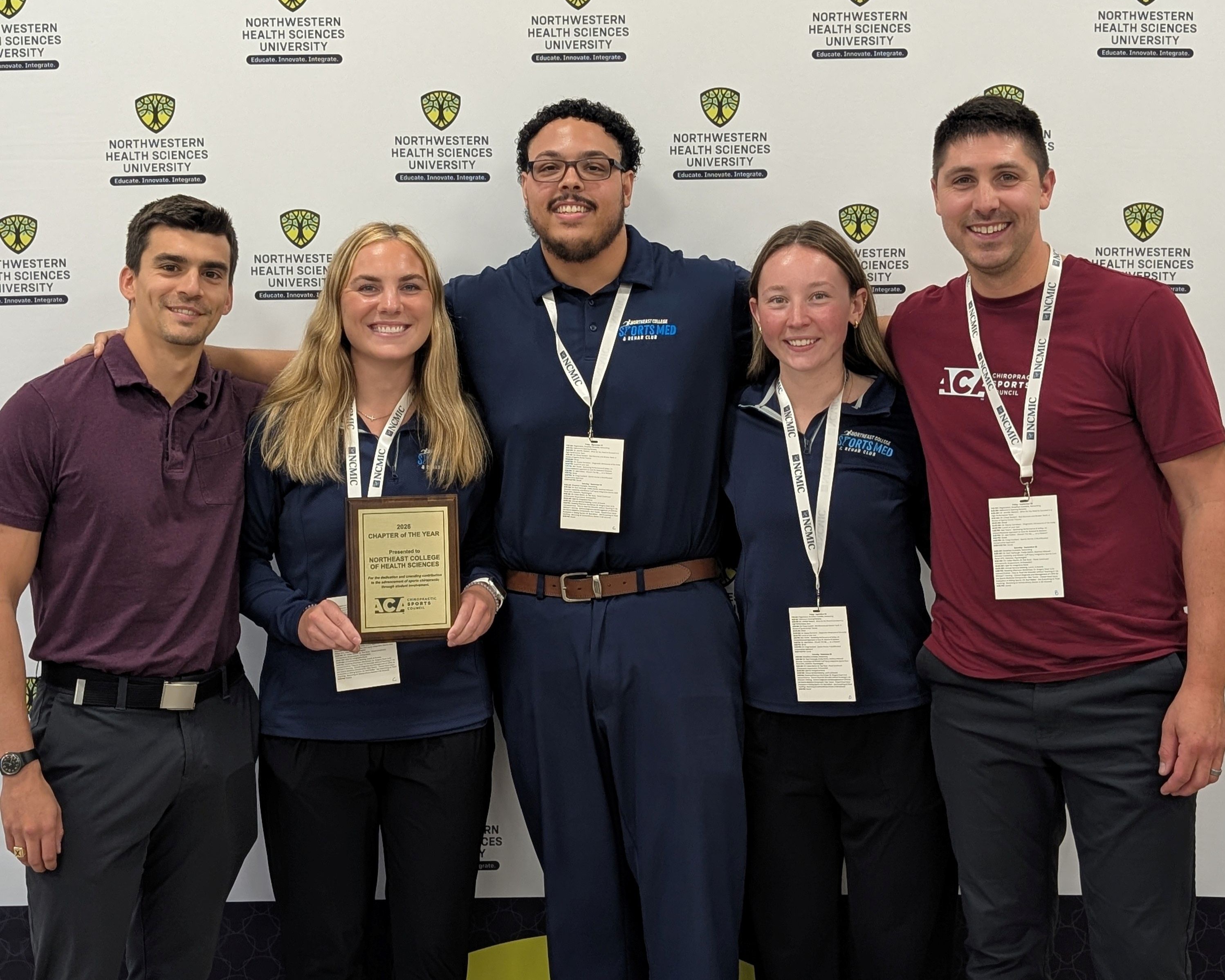 Five people holding a “Chapter of the Year” plaque awarded to Northeast College of Health Sciences, in front of a Northwestern Health Sciences University backdrop.