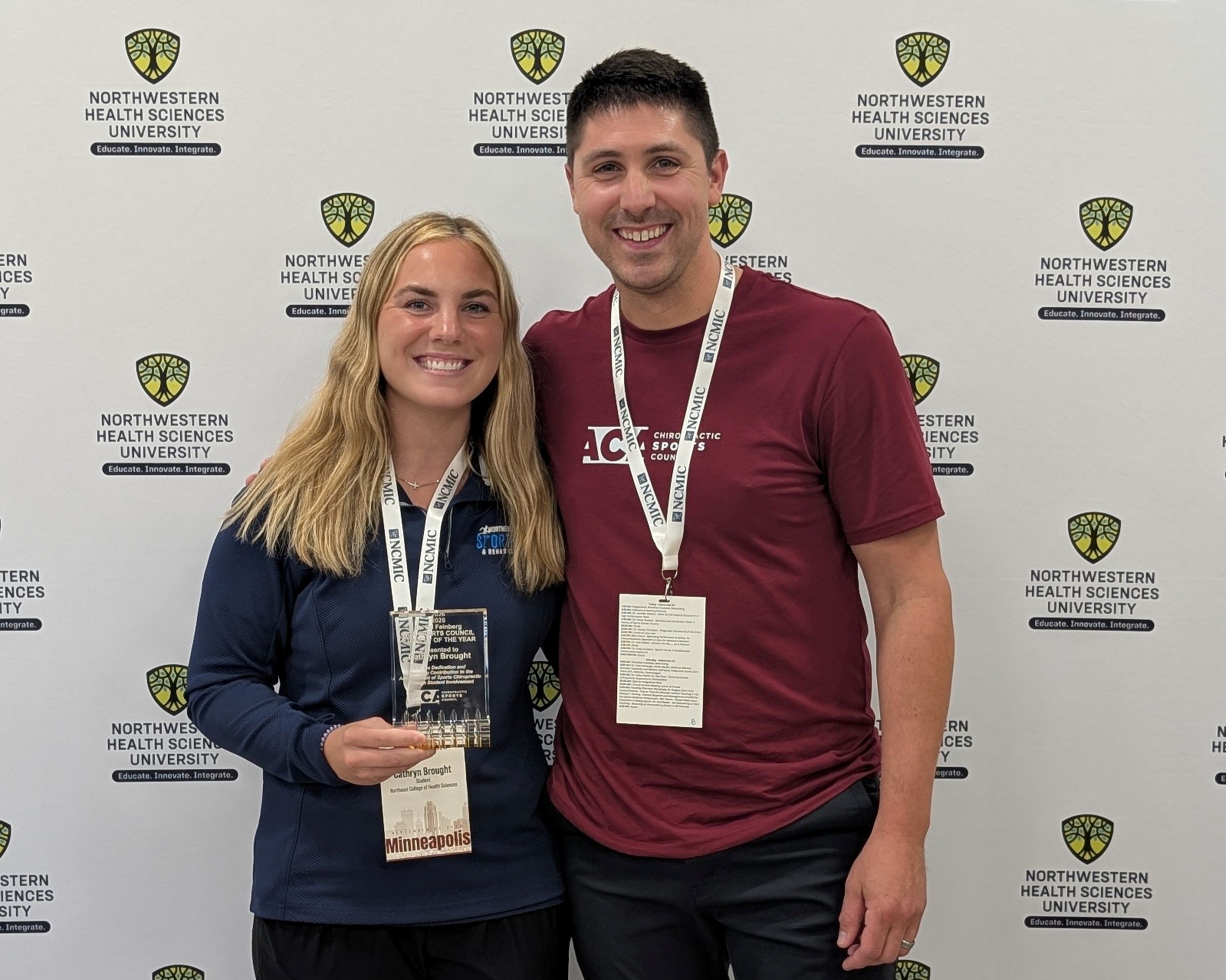 Two people posing with an award plaque in front of a Northwestern Health Sciences University backdrop at a conference.