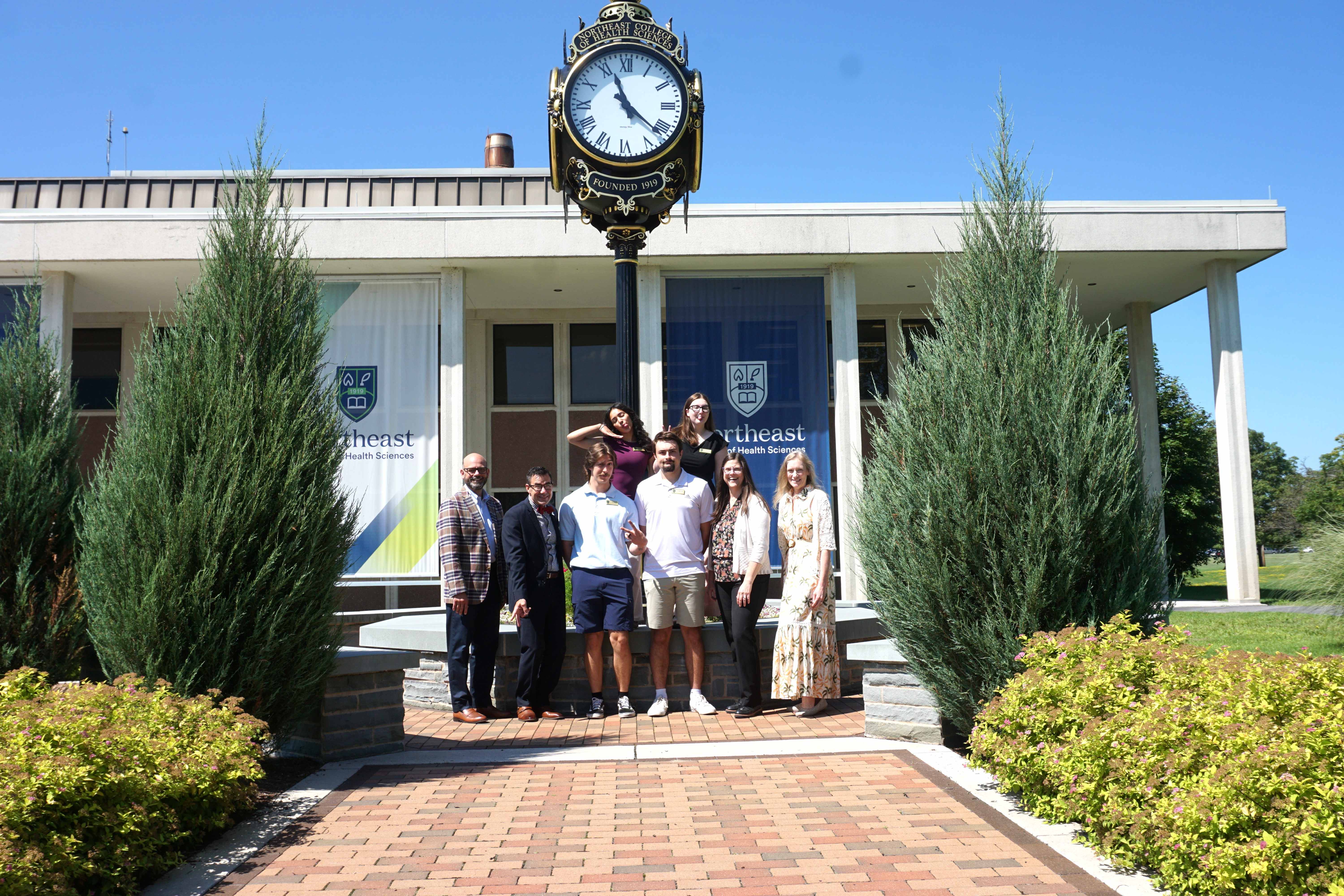 A group of students and staff posing in front of the Northeast College of Health Sciences clock tower and campus banners on a sunny day.