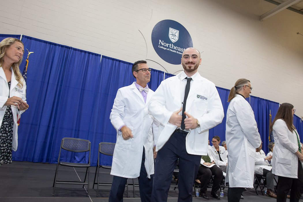 A student proudly showing off his new white coat at a Northeast College of Health Sciences white coat ceremony, with faculty and fellow students in the background.