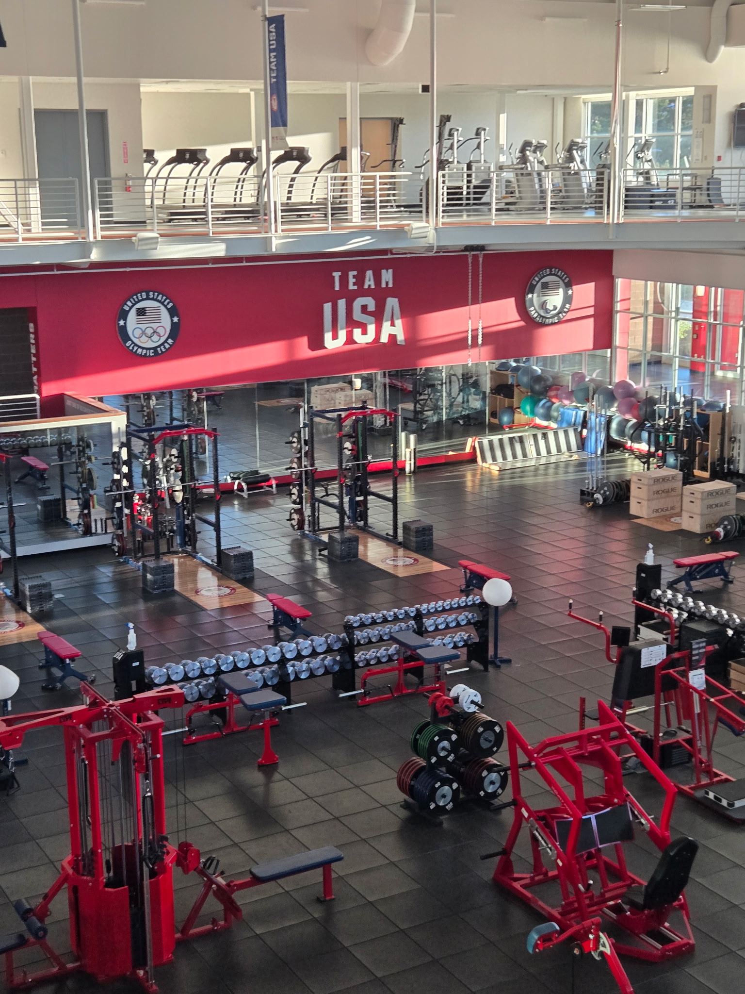 Interior view of the Team USA Olympic and Paralympic training center weight room, with red equipment, dumbbells, and the “Team USA” logo prominently displayed on a red wall.