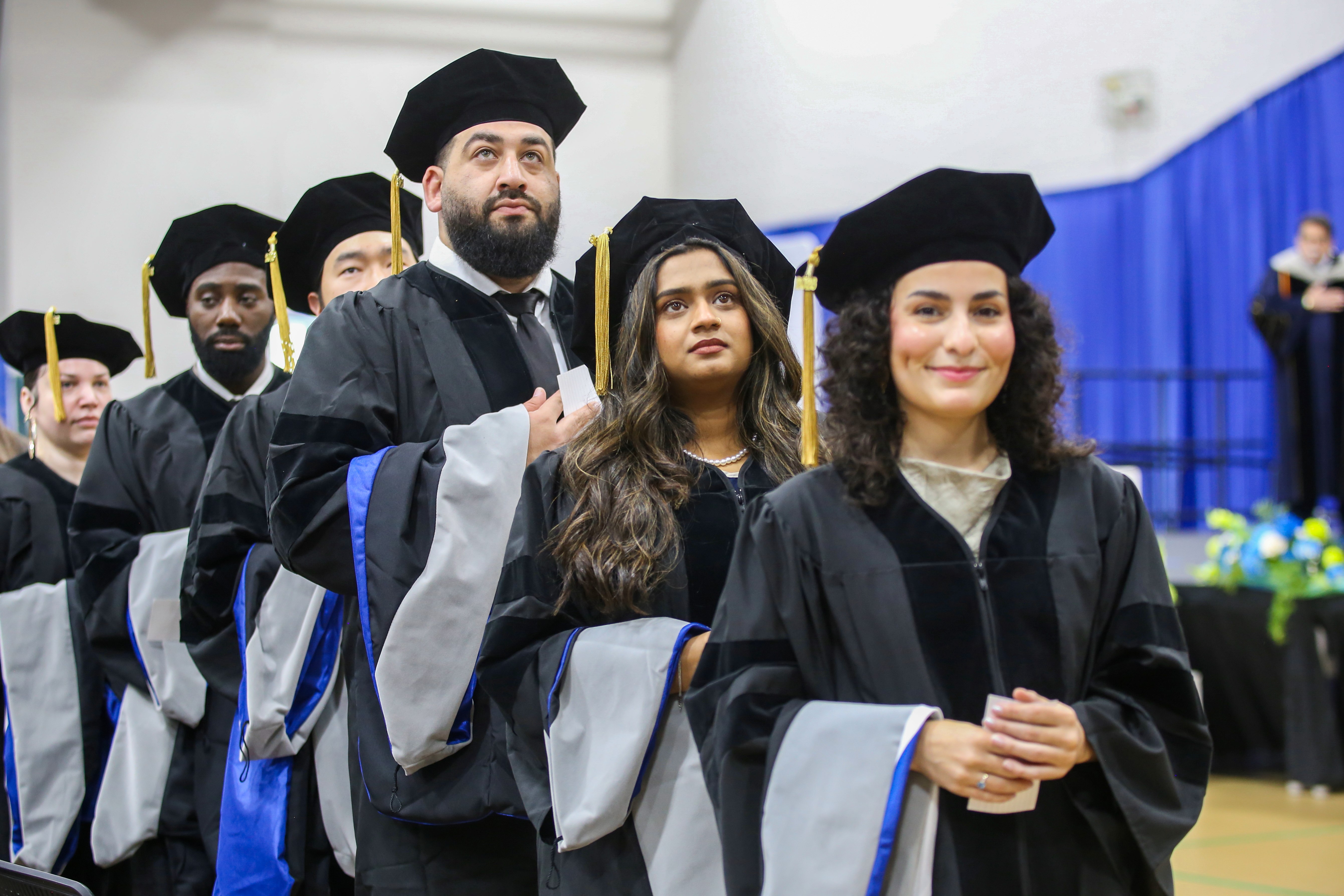 A row of graduates in black caps, gowns, and blue-trimmed hoods processing at a Northeast College of Health Sciences commencement ceremony.
