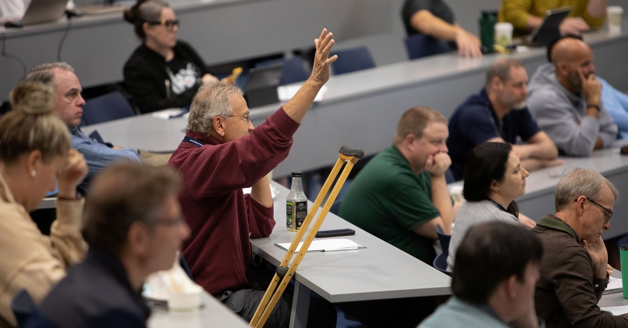 A man in a red sweatshirt raises his hand to ask a question during a lecture in a tiered classroom filled with attendees.