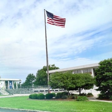 An American flag flying on a tall flagpole in front of a modern institutional building, surrounded by manicured green lawns, shrubs, and trees under a partly cloudy sky.