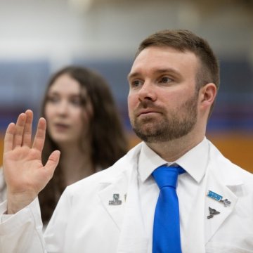 A man in a white coat and blue tie raises his right hand, appearing to take an oath at a Northeast College ceremony.