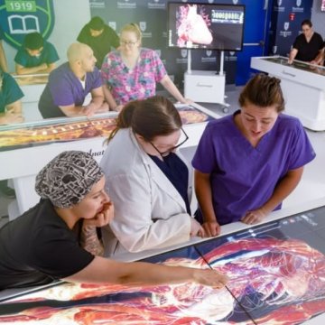 Students and an instructor in scrubs gathered around illuminated anatomy tables displaying detailed digital body scans in what appears to be a modern medical education lab known as an anatomage table..