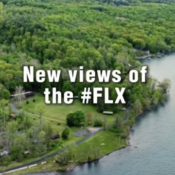 Aerial drone view of a lake shoreline surrounded by lush green forest, with text reading "New views of the #FLX."