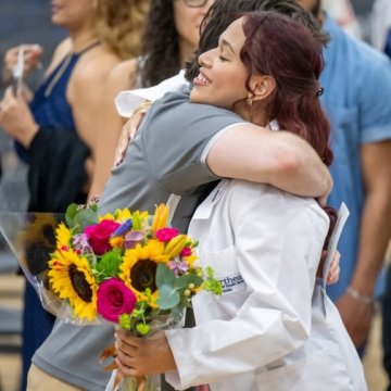 A graduate in a white coat hugging a loved one and holding a bouquet of sunflowers and pink flowers at a graduation ceremony.