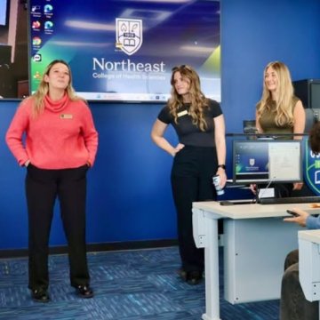 Three women in casual attire standing in front of a Northeast College of Health Sciences screen during a presentation.
