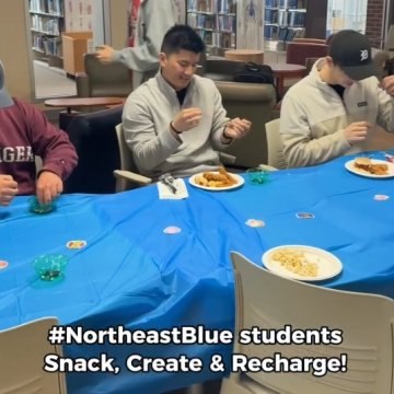 Three college students eating snacks at blue-covered tables in a library, at a "Snack, Create & Recharge" event for Northeast students