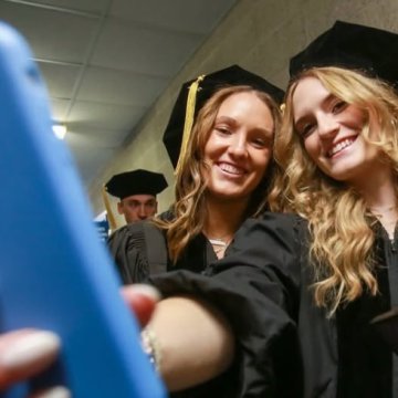 Two graduates in black caps and gowns smiling and taking a selfie before a commencement ceremony.