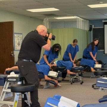 A photographer documenting chiropractic students in blue scrubs practicing adjustments on patients in a clinic lab setting.