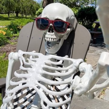 A plastic skeleton wearing red sunglasses relaxing in an Adirondack chair outdoors on a sunny summer day.