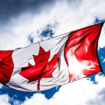 The Canadian flag waving against a dramatic cloudy sky, backlit by sunlight.