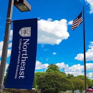 A Northeast College of Health Sciences banner on a lamppost with an American flag flying against a bright blue summer sky.