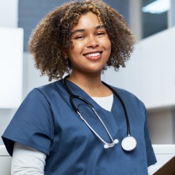 A smiling healthcare student or clinician in blue scrubs with a stethoscope around her neck, holding a clipboard in a bright medical setting.