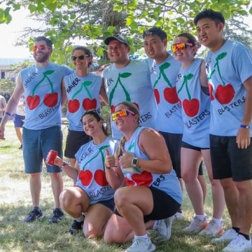 A group of eight people posing outdoors in matching light blue "Bauer Blasters" cherry-themed team shirts at what appears to be a summer event or field day.