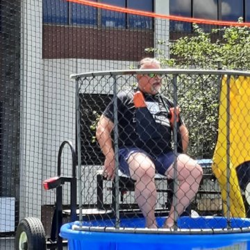 A man sitting on the platform of a dunk tank at an outdoor event, bracing himself above the water.