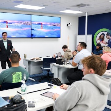 An instructor presenting to a group of students in a modern classroom with dual screens displaying a Zoom background of San Francisco.