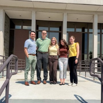 Five people smiling together outside the front entrance of a campus building on a sunny day.