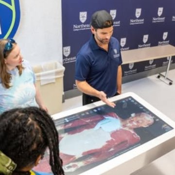 A Northeast College staff member demonstrating the digital anatomy table to students in front of a branded backdrop.