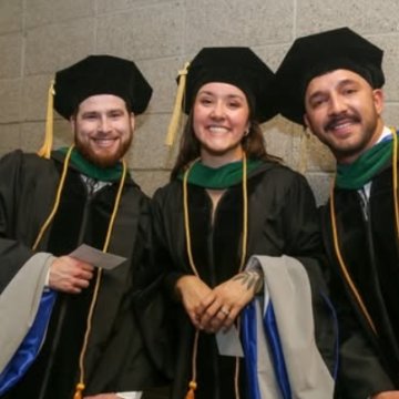 Three graduates in black doctoral caps and gowns with green and gold trim smiling together at commencement.