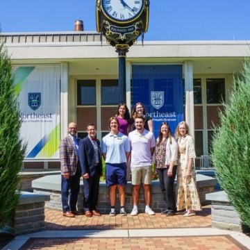 A group of students and staff posing in front of the campus clock tower and Northeast College of Health Sciences banners on a sunny day.