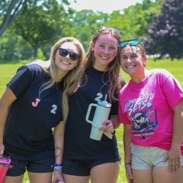 Three young women smiling together on a sunny green lawn at an outdoor campus event.