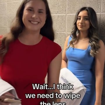 Two women holding white coats in a hallway, with on-screen text reading "Wait…I think we need to wipe the lens."