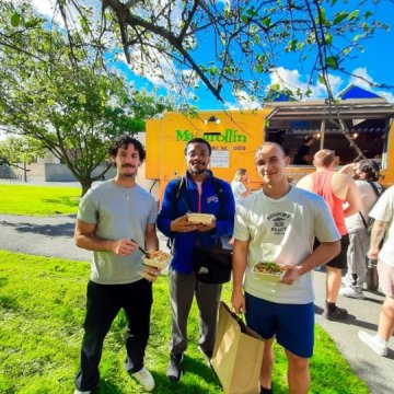 Three students smiling and holding food containers in front of a yellow "Macarollin" mac and cheese food truck on a sunny campus lawn.