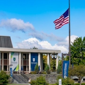 The Northeast College of Health Sciences main building with branded banners and the campus clock tower, with an American flag flying out front under a blue sky.