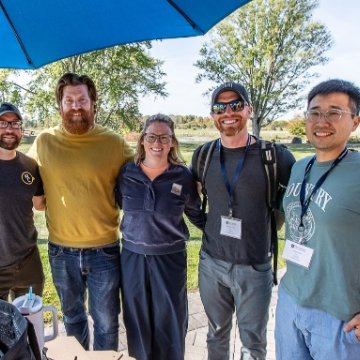 Five people smiling together under a blue umbrella at an outdoor campus event with fall foliage in the background.