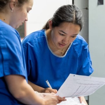 Two students in blue scrubs reviewing paperwork together in a hallway.