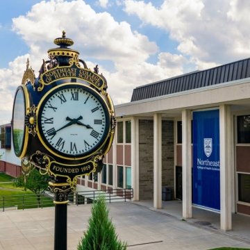 The Northeast College of Health Sciences ornate clock tower reading "Founded 1919" beside a campus building with a branded banner.