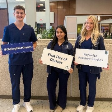 Three Physician Assistant Studies students in navy scrubs holding signs reading “#GoingNortheast,” “First Day of Classes,” and “Physician Assistant Studies.”