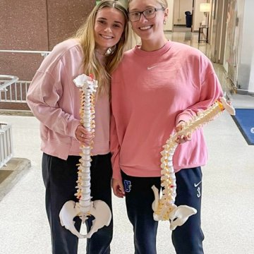 Two students in matching pink sweatshirts smiling in a campus hallway, each holding a spine and pelvis anatomical model.