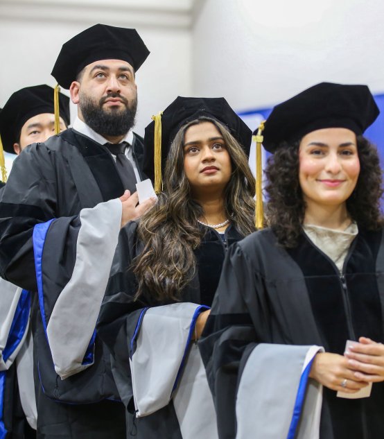 A row of graduates in black caps, gowns, and blue-trimmed hoods processing at a Northeast College of Health Sciences commencement ceremony.
