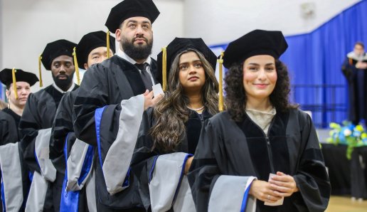 A row of graduates in black caps, gowns, and blue-trimmed hoods processing at a Northeast College of Health Sciences commencement ceremony.