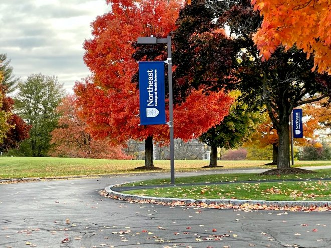 Northeast College of Health Sciences campus entrance in autumn, with blue branded banners lining a driveway surrounded by vibrant red and orange fall foliage.