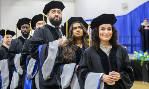 A row of graduates in black caps, gowns, and blue-trimmed hoods processing at a Northeast College of Health Sciences commencement ceremony.